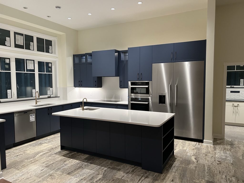 A modern kitchen with dark blue cabinetry and white countertops with plenty of windows and stainless steel appliances.