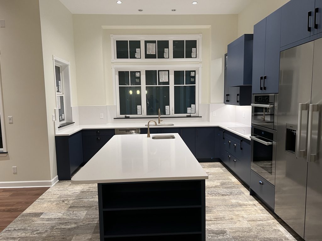 A modern kitchen with dark blue cabinetry and white countertops with plenty of windows and stainless steel appliances.