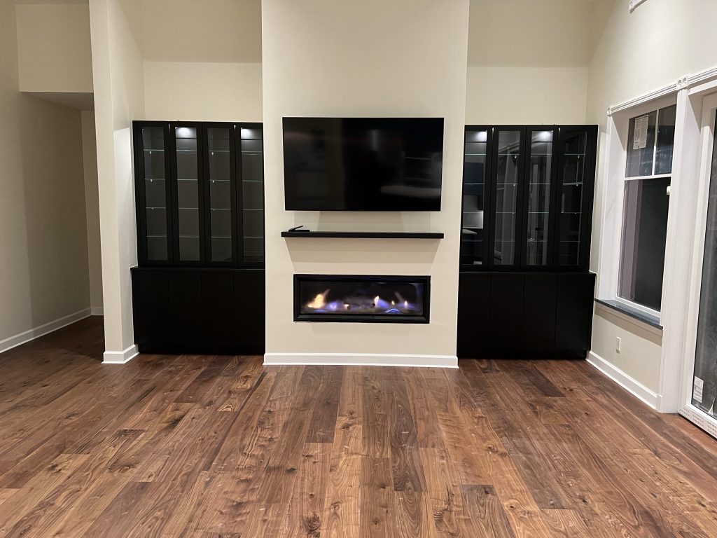 An empty living room with wood floors, a fireplace with a tv above it, and large dark cabinetry on either side.